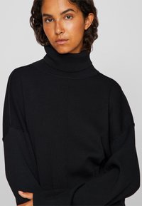 Μαύρο μπλουζάκι τύπου τερλίκι από ριγέ ύφασμα, με χαλαρή εφαρμογή, φαρδιά μανίκια και ψηλό γιακά για ζεστασιά.