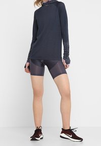 Sportovní oblečení s tmavým dlouhým rukávem s vzorovanými manžetami, kombinované s lesklými tmavými kraťasy a černými běžeckými botami.