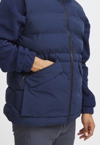Mornarska puffer jakna od flisa, s detaljima na zatvorenim džepovima i elastičnim manžetama. Ima kapuljaču i prednje zatvaranje na patentni zatvarač.