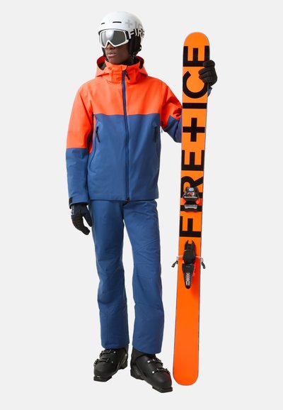 Lyžařský outfit s oranžovou a modrou bundou, ladícími kalhotami, černými rukavicemi a bílou helmou. Drží oranžovou lyži s černým nápisem.