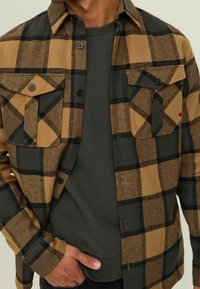 Uomo che indossa una camicia a quadri marrone e nera sbottonata sopra una maglietta a maniche corte grigia scura, con una mano infilata nella tasca dei pantaloni.