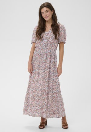 Giovane donna che indossa un lungo abito floreale chiaro con maniche a sbuffo e sandali marroni con tacco, in piedi davanti a uno sfondo semplice.