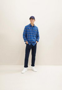 Niebieska koszula w kratę z dwiema kieszeniami na piersiach, granatowe spodnie i białe trampki. Model w granatowej czapce, stojący na neutralnym tle.