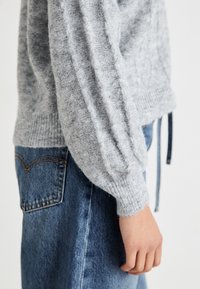 Pull en tricot gris texturé avec des poignets côtelés, offrant une coupe décontractée. Assortie à un jean en denim bleu mettant en avant des détails de couture visibles.