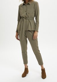 Olivgrünes, strukturiertes Outfit mit tailliertem Gürtel, bestehend aus einem Button-Up-Hemd mit zwei Brusttaschen und slim-fit Hosen. Braune Wildleder-Stiefeletten.