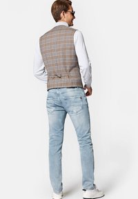 Brązowa kamizelka w kratę o dopasowanym kroju, w zestawie z jasnoniebieskimi dżinsami. Kamizelka ma zapięcie na plecach i wysoki dekolt, noszona z białą koszulą.