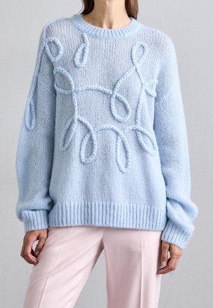 Pull tricoté bleu clair avec motifs en boucles en relief porté avec un pantalon rose clair, montré du cou aux cuisses sur fond uni.