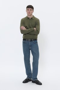 Camisa polo de manga larga verde con cuello, combinada con vaqueros azules holgados y zapatos de vestir negros. Diseño simple y casual, con acentos mínimos.