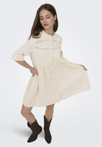 Fiatal nő, hosszú barna hajjal, krémszínű gombos ruhát csipkés részletekkel és sötétbarna bokacsizmát visel, csukott szemmel pózol.