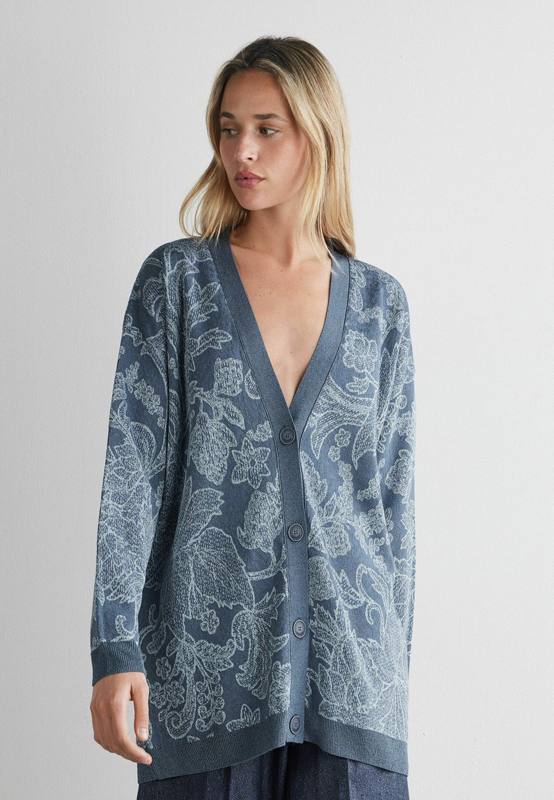 Blond vrouw die een oversized blauwe cardigan met knopen en een licht bloemenpatroon draagt, staand tegen een effen witte achtergrond.