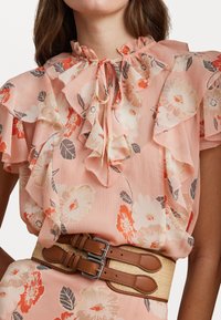 Blomstret bluse i blødt ferskenfarvet stof med flæsedetaljer, knyttet halsudskæring og livlige orange og cremefarvede blomster. Parret med et beige bælte.