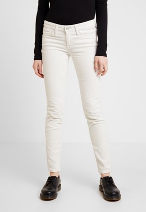 Person wearing off-white slim-fit corduroy pants, black long-sleeve top, and black lace-up shoes standing against a plain background.