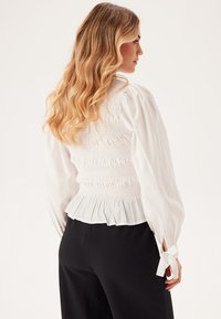 Hvid smocket bluse med pufærmer, plissert kant og bånddetaljer ved manchetterne. Blød stof med teksturerede detaljer. Sorte bukser er ikke synlige.