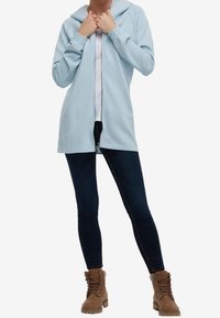 Sudadera con cremallera de color azul claro, con capucha, hecha de un material suave. Llevada sobre una blusa blanca y jeans oscuros, combinada con botas marrones de cordones.