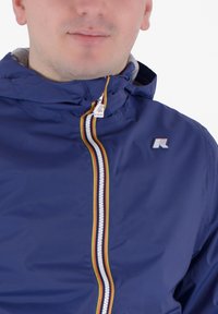 Námornícky vetrovka s kapucňou, obsahujúca biely zips s viacfarebnými pruhmi a malým logom na hrudi. Hladký povrch.