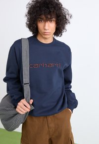 Navy blue sweatshirt with ribbed crew neck, featuring "carhartt" in brown embroidery. Paired with brown shorts and a gray plaid bag.