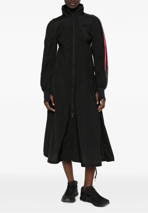 Persona che indossa un vestito nero a maniche lunghe con zip, colletto alto e una striscia rossa su una manica, abbinato a scarpe sportive nere.