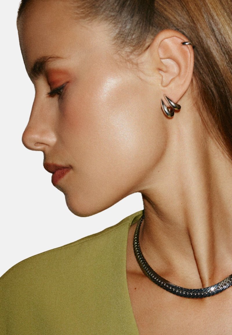 Silver spiralformade örhängen pryder örat, kombinerade med ett metalliskt halsband. Bakgrunden är enkel vit och framhäver smyckena.