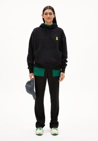 Felpa nera con logo a nastro giallo, maglietta verde a strati, pantaloni neri, cappellino grigio in mano e sneakers bianco-verdi.