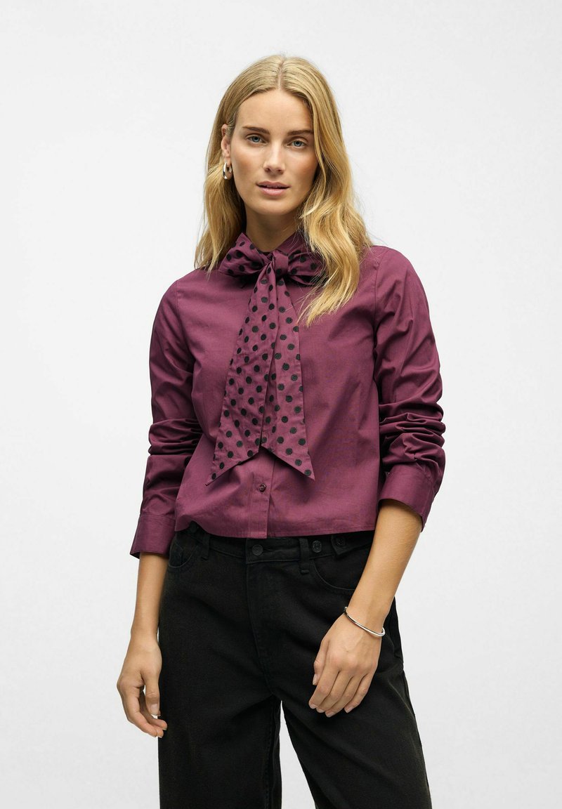 Vinrød knap-op skjorte med stor polkaprikket butterfly, med lange ærmer og en glat tekstur, kombineret med sorte bukser.