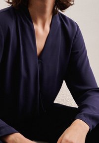 Blouse marine avec un col en V et des manches longues, fabriquée dans un tissu lisse, présentant un détail de bouton unique au niveau du col. Fond texturé.