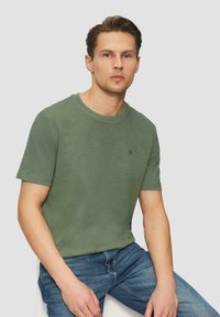 T-shirt z krótkim rękawem w stonowanym zielonym kolorze, wykonany z bawełny, z dyskretnym logo i luźnym krojem, w połączeniu z niebieskimi dżinsami.