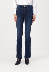 Jean évasé bleu foncé en denim. Comprend cinq poches, un système de fermeture par bouton et des coutures contrastées. Associé à des bottines noires.