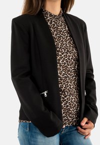 Femme portant un blazer noir avec des poches zippées sur un haut à imprimé léopard et un jean bleu, les mains reposant près de la taille.