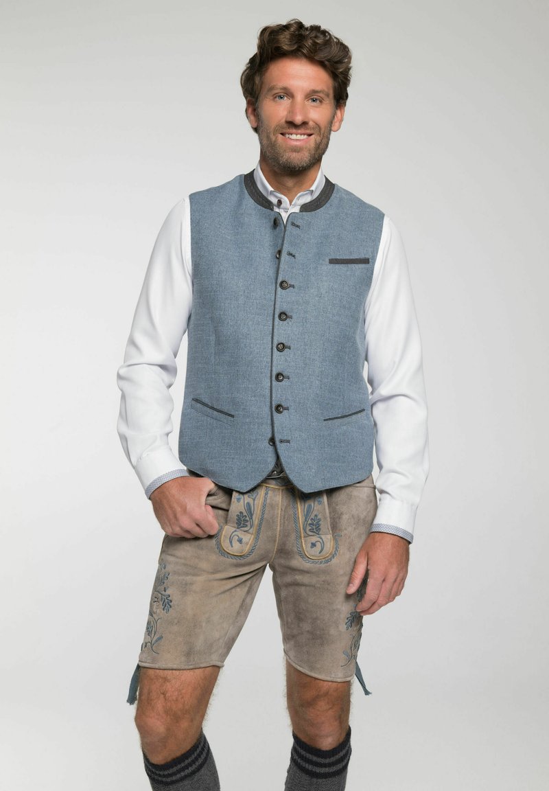 Blauer Weste mit schwarzen Akzenten, kombiniert mit beigen Lederhosen mit floralen Stickereien. Graue knielange Socken runden das Outfit ab.