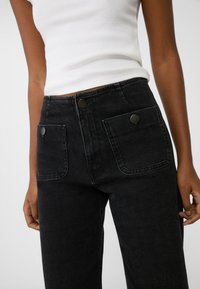 Pantalones de mezclilla negros de tiro alto con corte de pierna recta, dos bolsillos delanteros y costuras en contraste. Presentan detalles de botones metálicos.