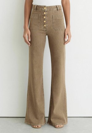 Brune bukser med høj talje og brede ben, med fem knapper foran, to store forlommer, båret med en hvid top og sandaler.