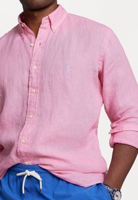 Ljust rosa linneknappad skjorta med en liten blå logotyp på bröstet, uppvikta ärmar, kombinerad med blå shorts med dragsko.