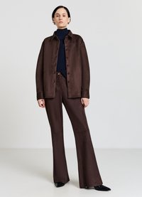 Braune Satinjacke mit Knopfverschluss und Hemdkragen, kombiniert mit braunen Hosen mit weitem Bein. Das Modell trägt darunter einen marineblauen Rollkragenpullover.