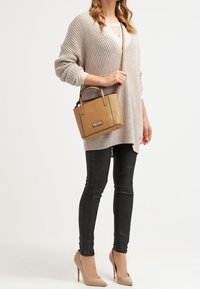 Sac à main beige avec un design structuré et deux poignées. Porté avec un pull en maille surdimensionné beige clair et un jean skinny noir, complété par des talons couleur nude.
