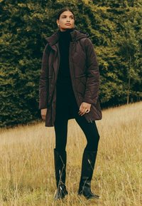 Pruun tepitud jakk kapuutsiga, kantud musta kõrge kaelusega kampsuni ja kitsaste mustade pükste peal. Komplekti täiendavad kõrged mustad saapad. Taustal on rohumaa.