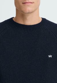 Temno modra pletena majica z rebrasto teksturo, krojem okoli vratu in belim logotipom na prsih.