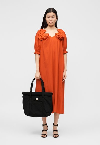 Γυναίκα με πορτοκαλί μίντι φόρεμα με βολάν στους ώμους κρατά μια μεγάλη μαύρη τσάντα tote και φορά μαύρα σανδάλια με λουράκια.