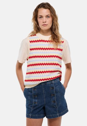 Femme aux cheveux ondulés portant un gilet en maille rayé sur une chemise beige à manches courtes et un short en jean taille haute avec des boutons.