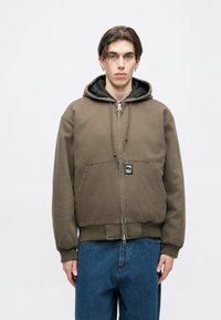 Smeđa bomber jakna s kapuljačom, s prednjim džepovima, rebrastim manžetama i rubom. Ima crnu unutrašnju postavu i logo na prednjem dijelu.