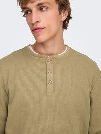 Chemise beige en maille gaufrée à manches longues avec un col arrondi, une patte de boutonnage à trois boutons et une surface texturée.