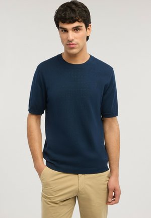 Jeune homme aux cheveux courts, bouclés et foncés, portant un polo en maille bleu marine à manches courtes et un pantalon beige, debout avec une main dans la poche.