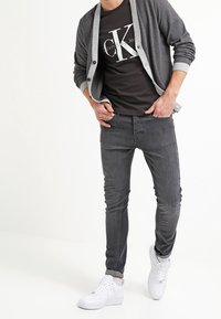 Grauer Cardigan mit hellgrauen Akzenten, schwarzes T-Shirt mit weißem Logo, graue Jeans und weiße Turnschuhe. Lässiges geschichtetes Outfit.