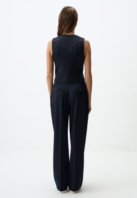 Marineblaues ärmelloses Top, kombiniert mit einer weit geschnittenen Hose. Die Hose hat zwei Gesäßtaschen mit Knopfdetails. Glatter, strukturierter Stoff.