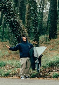 Un jeune homme portant un hoodie bleu marine, un pantalon beige et des baskets noires, tenant une planche de surf et une veste, se tenant parmi un feuillage vert et des arbres.