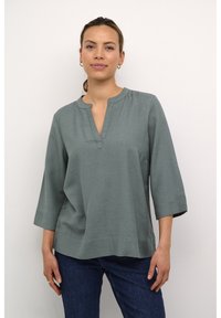 Blusa di colore verde chiaro, morbida e con una vestibilità ampia, scollo tondo e maniche a tre quarti. Texture liscia, dettagli minimi e forma rilassata.
