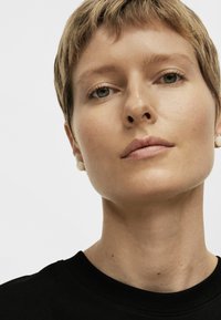 Primer plano de una persona con cabello corto y castaño claro que lleva pendientes redondos pequeños. Lleva una camiseta negra con cuello redondo. La piel es suave.