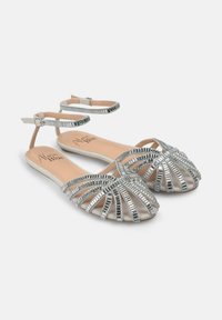 Sandali piatti argento con design a fascia incrociata, adornati con accenti metallici. La suola è di un beige chiaro e le fasce hanno una superficie liscia.