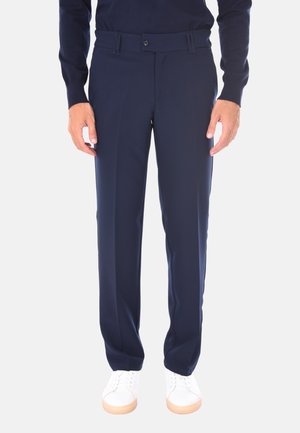 Uomo che indossa pantaloni su misura blu navy, maglione blu navy e sneaker bianche, in piedi con le braccia rilassate ai lati su uno sfondo bianco.