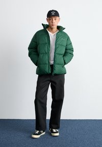 Giacca a vento verde con zip frontale e colletto alto, indossata su una t-shirt bianca, abbinata a pantaloni neri e sneaker verdi.