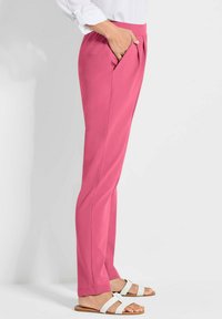Getailleerde, hooggewaisting roze broek van zachte stof, met zijzakken en een rechte pijp. Draagt witte sandalen.